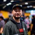 黄油游戏库社区运营总监张博文的职业照，男性，戴棒球帽，穿休闲卫衣，背景为玩家社区活动现场